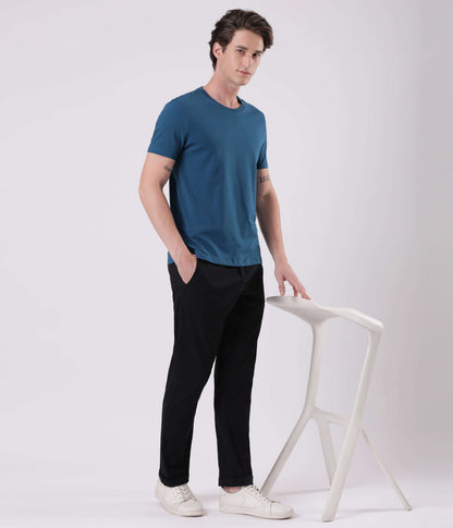 Model wearing Turms Black Sea Chinos in black paired with a teal t-shirt and white sneakers, standing by a minimalist stool.