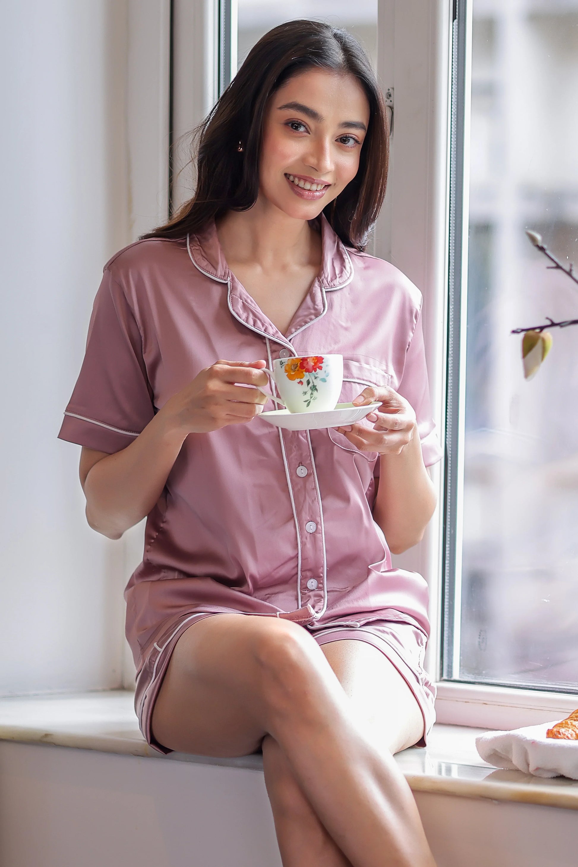 Woman in pink shorts set holding a floral teacup by a window
